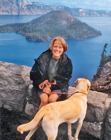 Photo of Amy Wroe Bechtel smiling in front of a lake while holding the leash of a yellow Labrador retriever