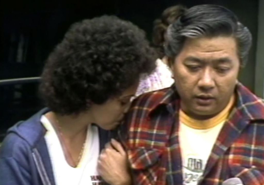 A crying woman grips her husband's arm and presses her face against his shoulder.