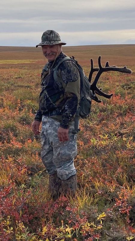 An older man dressed in hunter's camouflage stands smiling in an open field, carrying a set of antlers on his back.