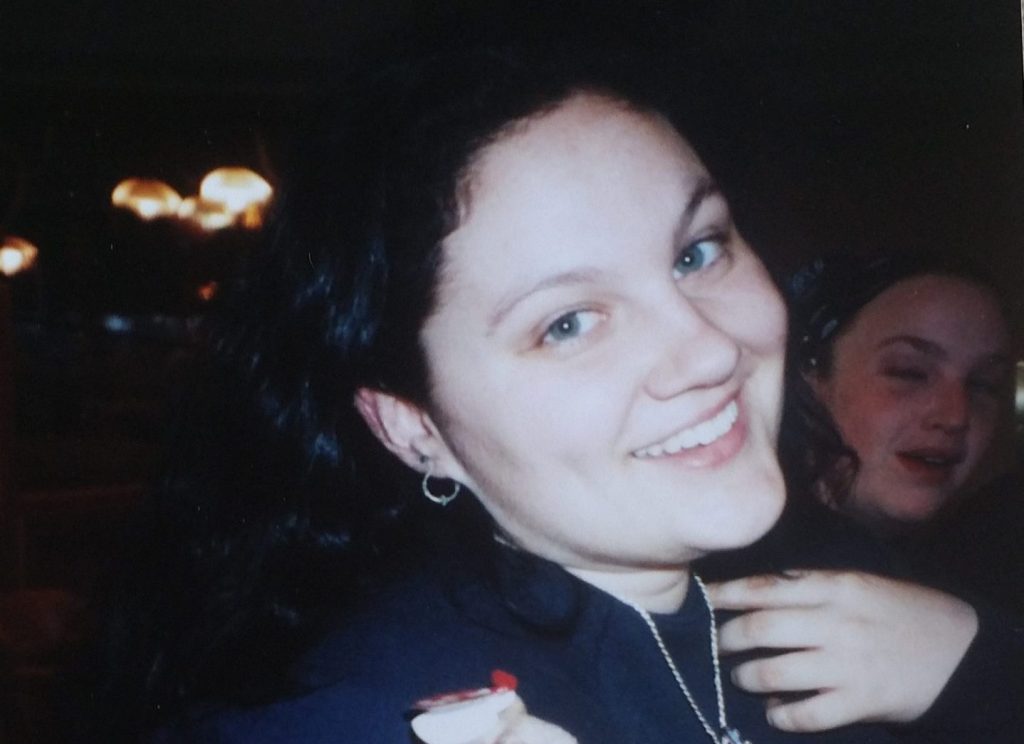Close-up of a smiling young woman with dark hair, wearing a black shirt and silver earrings, with another person partially visible in the background.