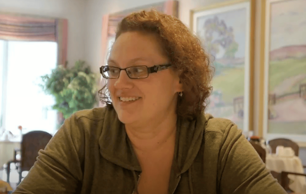 A smiling woman with curly hair and glasses sitting at a table in a cafe, with a soft-focused background featuring plants and artworks.